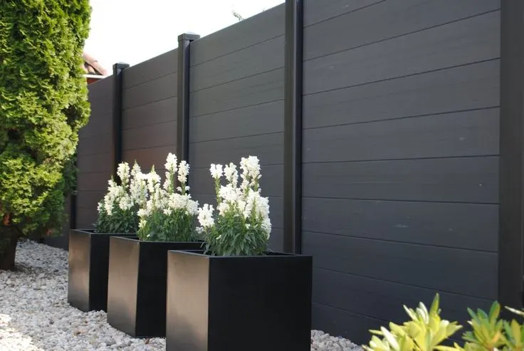 A dark grey aluminum fence with black planters containing white flowers in front of it