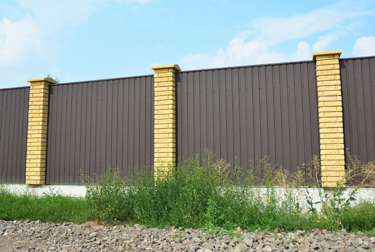 Brown corrugated metal fence with sandstone brick pillars beside a grassy verge