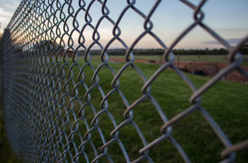 Close-up of a galvanised chain link fence running alongside a green field at sunset