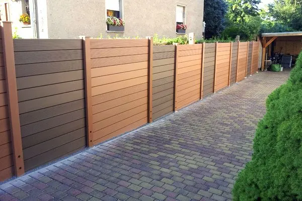 A long, straight fence made of composite panels in varying shades of brown, installed along a brick driveway