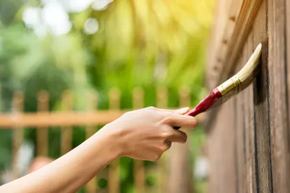 A person wearing a red glove painting a white wooden fence with a brush