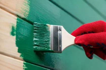 A gloved hand painting a wooden fence green with a brush