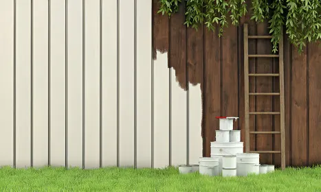 A wooden fence being painted with white paint, with a ladder leaning against it and paint cans on the grass in front