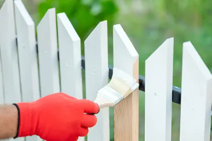 A person wearing a gardening glove painting a white wooden fence with a brush