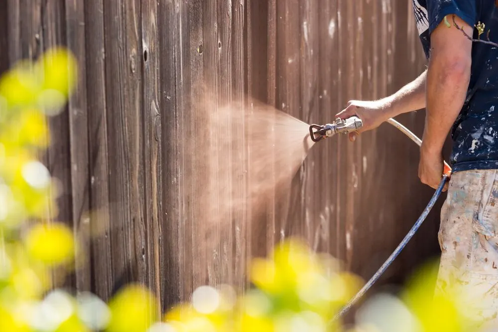A person using a spray gun to paint a wooden fence