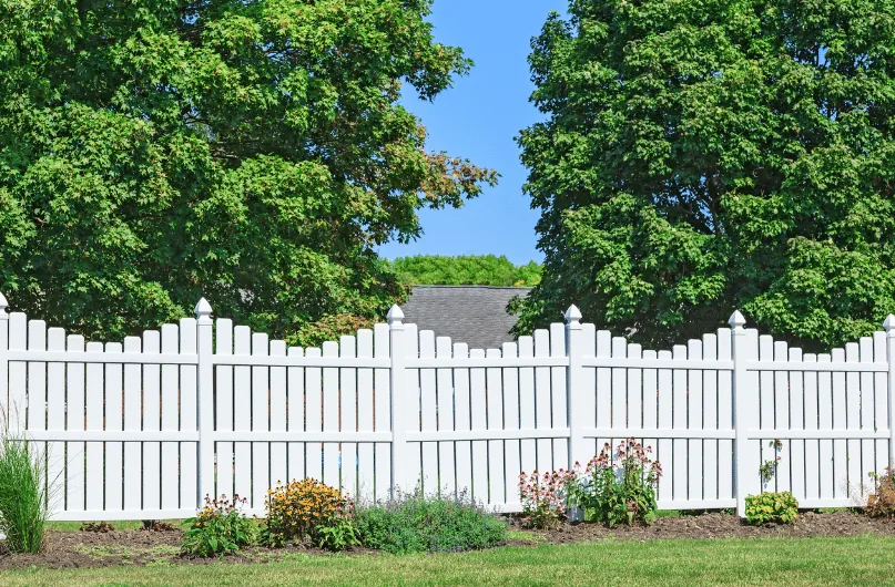 White vinyl picket fence in a garden setting with flower beds and mature trees behind