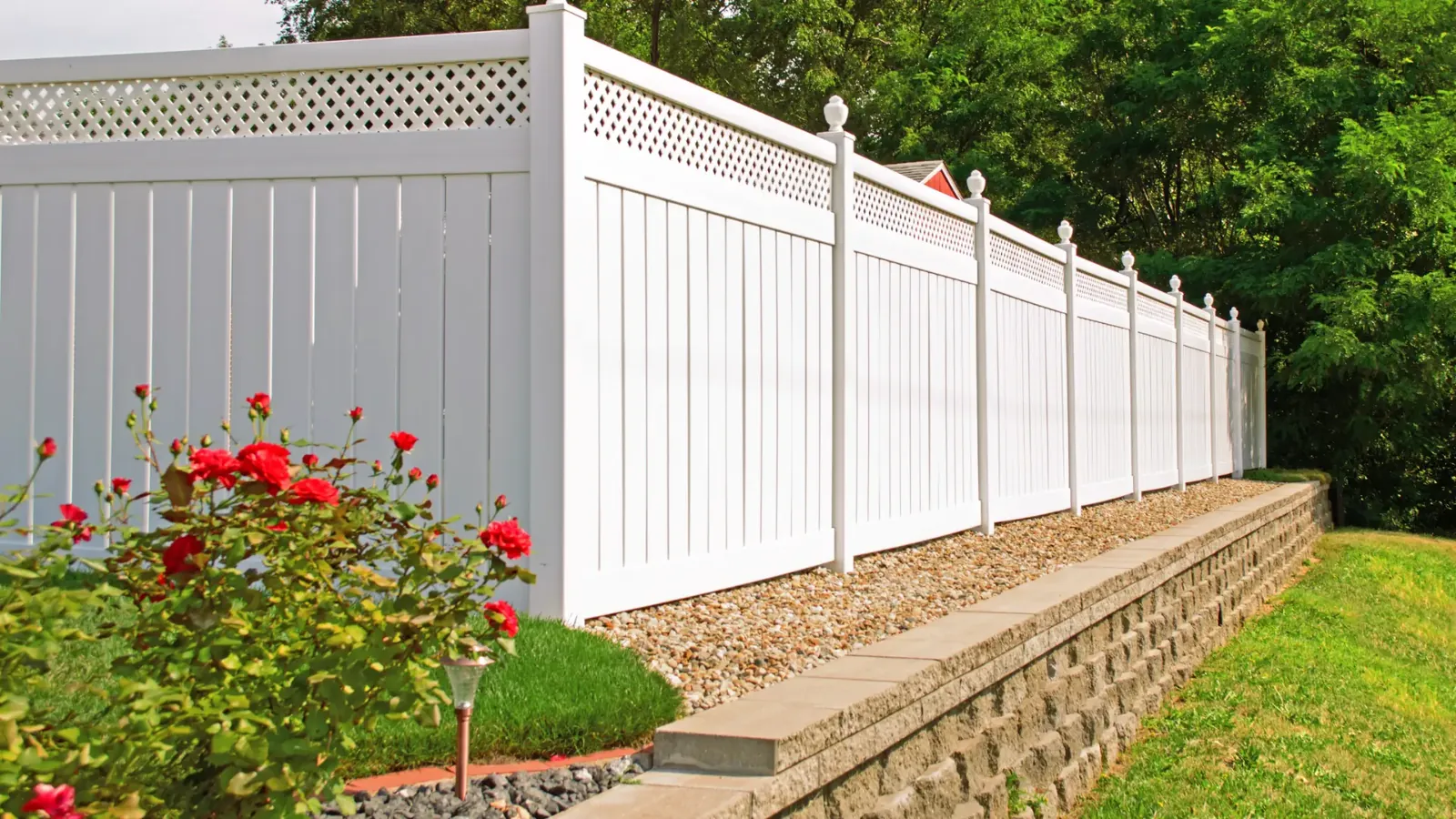 A white vinyl fence installed along a stone retaining wall in a garden setting